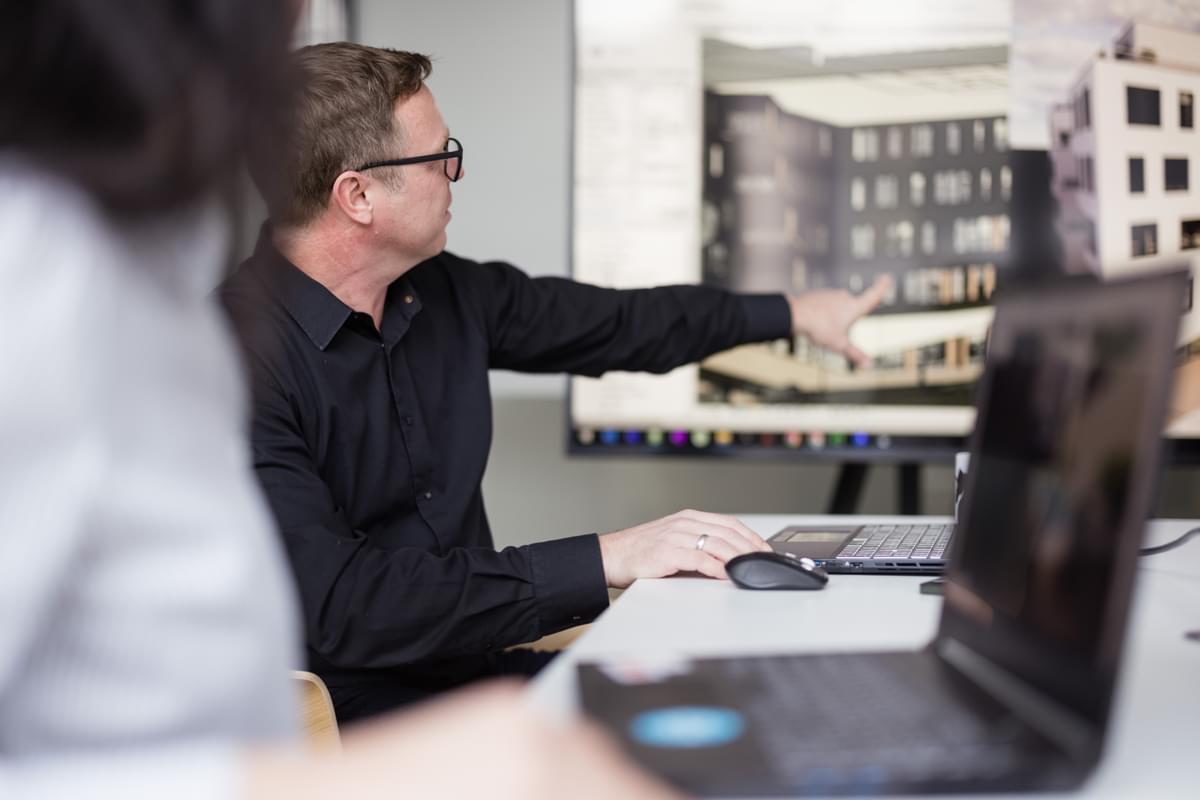 An architect in a design meeting, pointing to a 3D architectural visualization of a building on a large monitor.