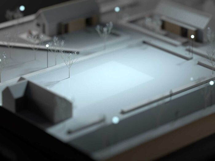 Architectural scale model with small buildings, pathways, and trees arranged around an open central space, illuminated by soft lighting